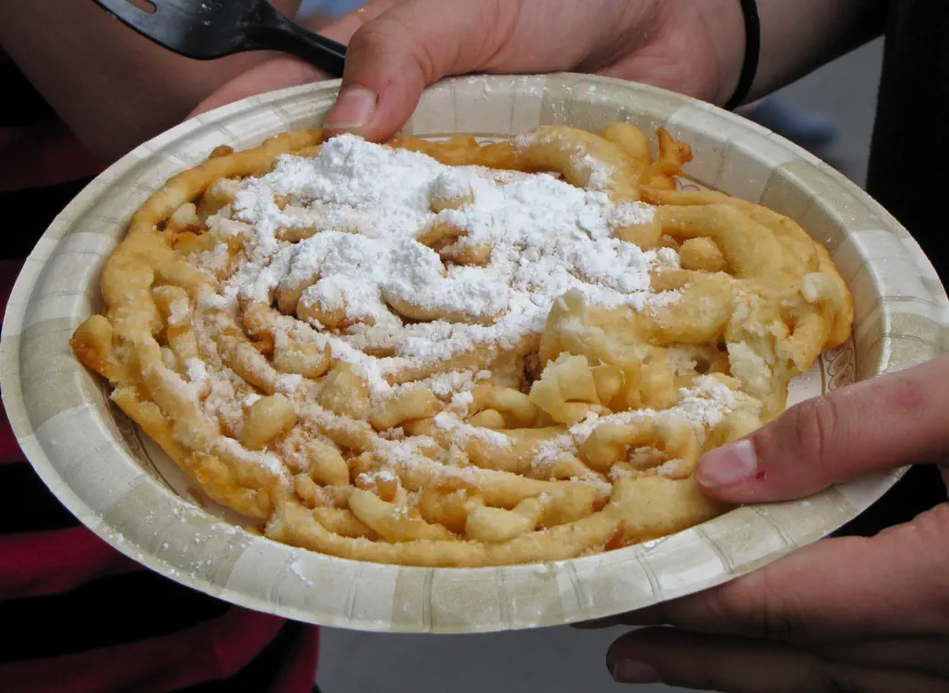 A funnel cake under powdered sugar, handed over on a paper plate